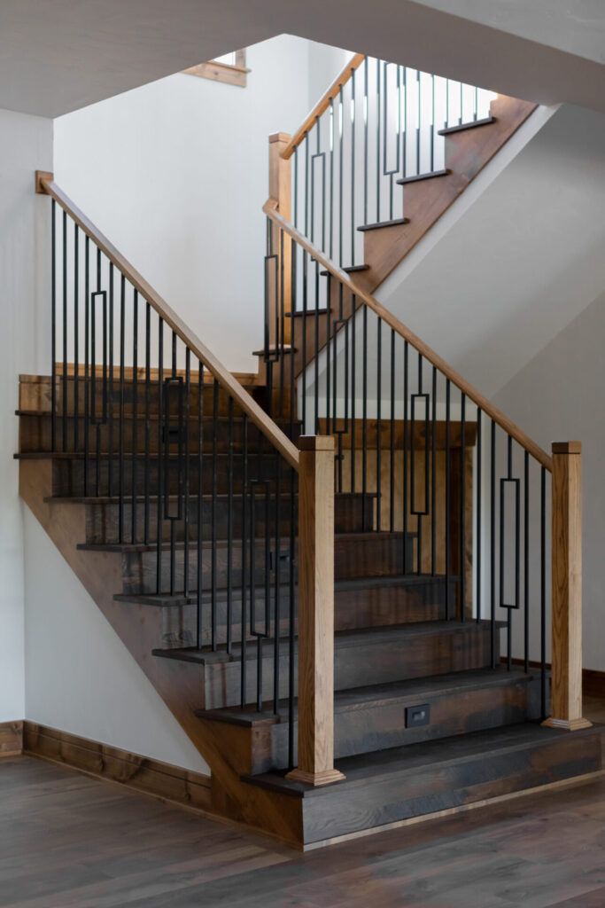 A modern staircase by Owen Custom Homes features dark wood steps and a black metal railing with a geometric design. Vertical bars with square and rectangular patterns complement the wooden handrails, matching the step finish. The space is beautifully illuminated by natural light.