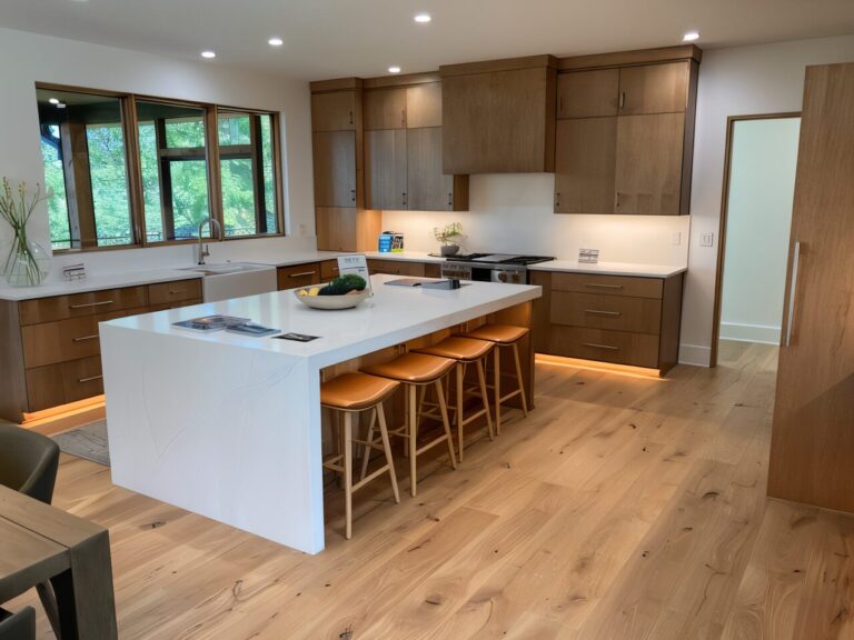 Modern kitchen with wooden cabinets, white countertops, and a large island with four orange barstools. Wide windows provide natural light. Custom homes by Richardson Homes showcase this clean, contemporary design with light wood flooring.