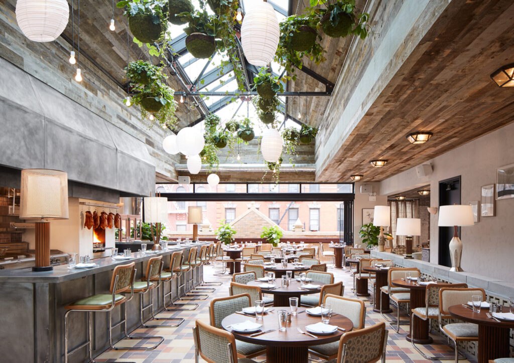 A modern restaurant with a high, partially glass ceiling, hanging plants, and round paper lanterns offers an inspiring starting point for design ideas. Enjoy city views from large windows, exposed concrete, a long bar with stools, and stylishly set tables.