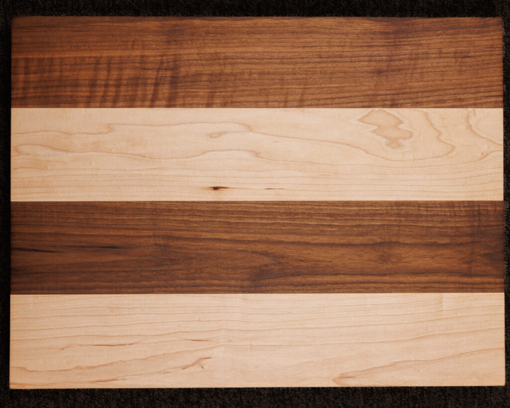 The 24 x 18 Custom Cutting Board features alternating horizontal bands of light and dark wood grain for a striking striped look, set against a dark, textured background.