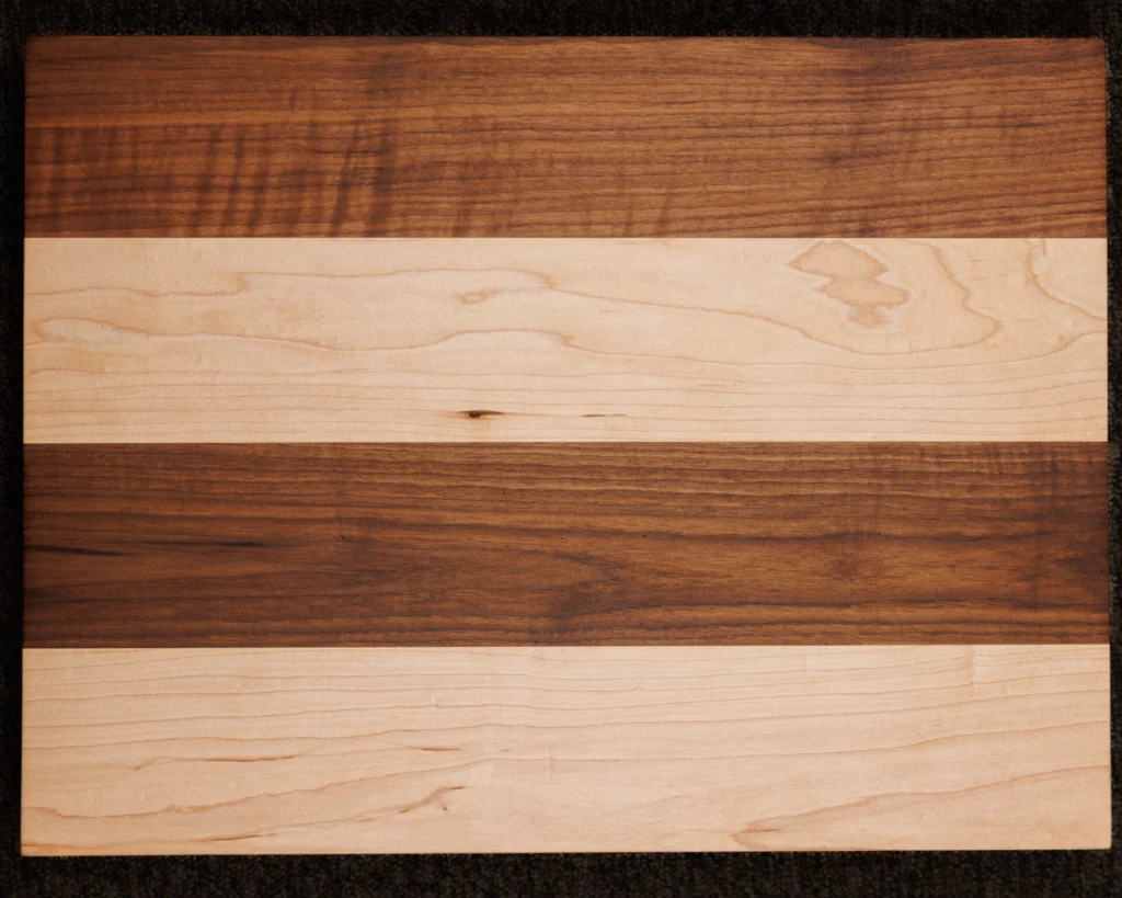 The 24 x 18 Custom Cutting Board features alternating horizontal bands of light and dark wood grain for a striking striped look, set against a dark, textured background.